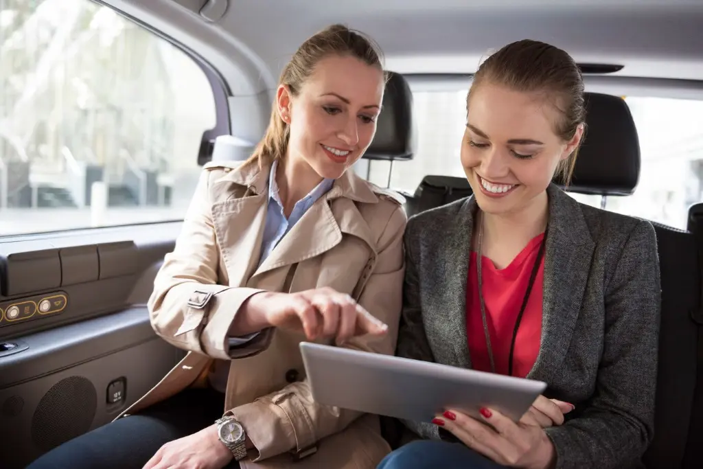 two young women customer and real estate agent talking back seat luxury car tablet look properties smilling