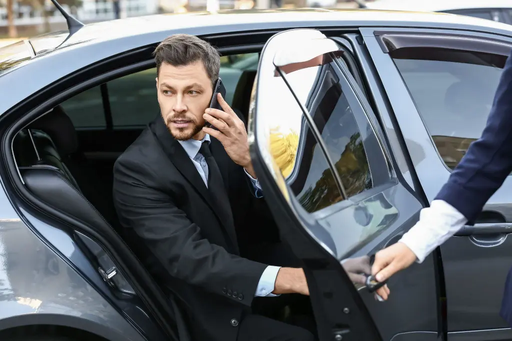 young businessman talking on smartphone getting out of luxury car opened by private chauffeur