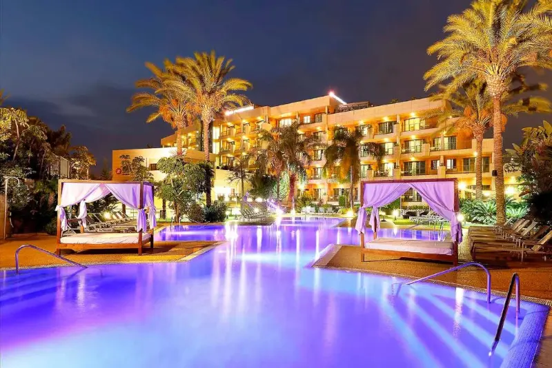 Vista frontal de noche de piscina y hotel Exe Estepona Thalasso Spa con palmeras y hamacas en Estepona